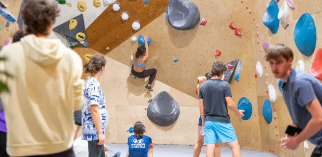 une grimpeuse sur un mur, les autres sur les tapis regardent.